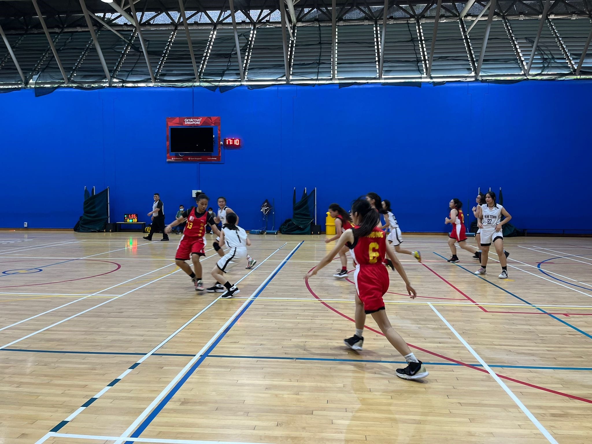 Girls in red and white uniforms play basketball on an indoor court with a blue wall and scoreboard showing 17:10.